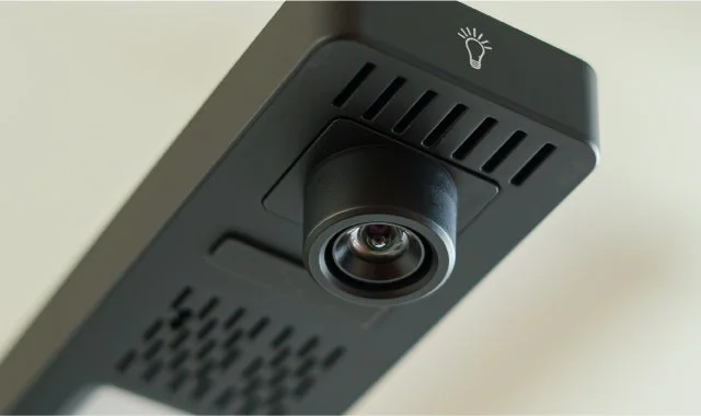 黒い電子機器のクローズアップ画像です。中央にレンズのような部品があり、上部には通気口と電球のアイコンが見えます。プロジェクターやスマートカメラなどのデバイスの一部と考えられます。